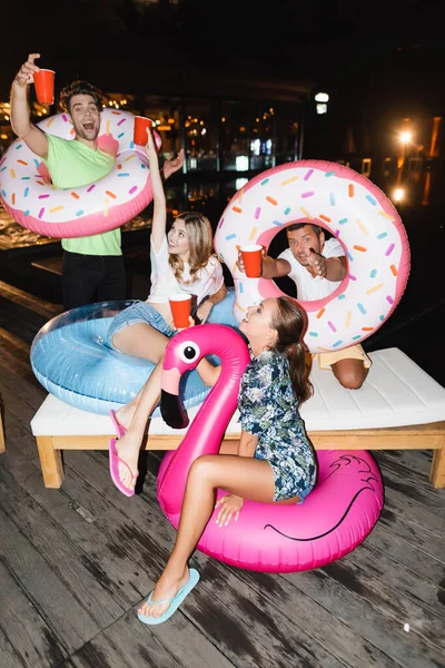Jeunes amis avec des anneaux de natation tenant des tasses jetables pendant la fête la nuit — Photo de stock