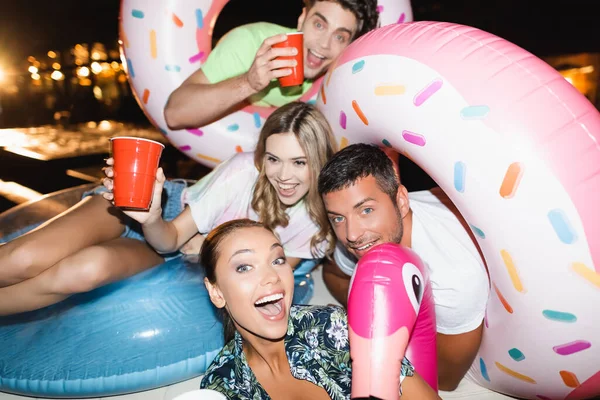 Enfoque selectivo de amigos emocionados con anillos de natación que sostienen tazas desechables durante la fiesta por la noche - foto de stock