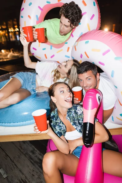 Selective focus of excited woman sitting on swim ring near boyfriend and friends with disposable cups at night — Stock Photo