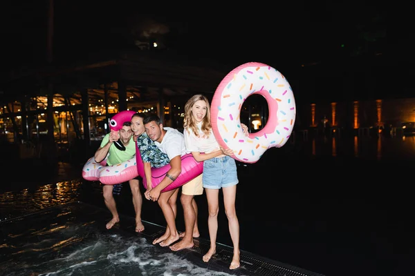 Excited young friends with swim rings looking at camera during party near pool at night — Stock Photo