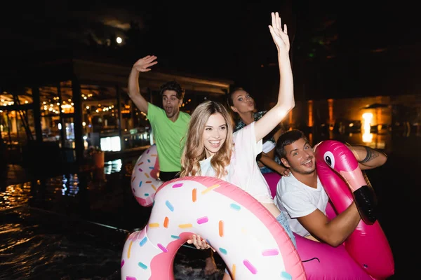 Выборочный фокус молодой женщины, держащей плавательное кольцо и машущей рукой рядом с друзьями во время вечеринки у бассейна ночью — стоковое фото