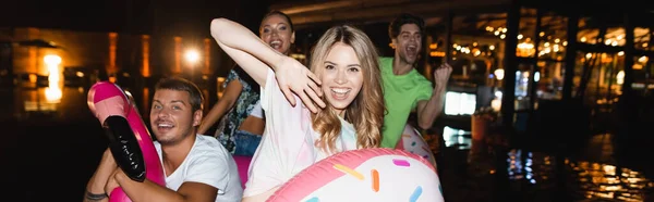 Sito web intestazione della donna con anello di nuoto guardando la fotocamera vicino agli amici durante la festa vicino alla piscina di notte — Foto stock