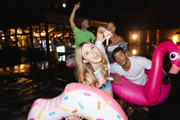 Selective focus of young woman showing peace near friends with swim rings during party at night — Stock Photo