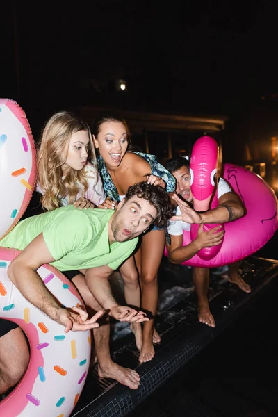 Enfoque selectivo de amigos con anillos de natación de pie cerca de la piscina durante la fiesta por la noche - foto de stock