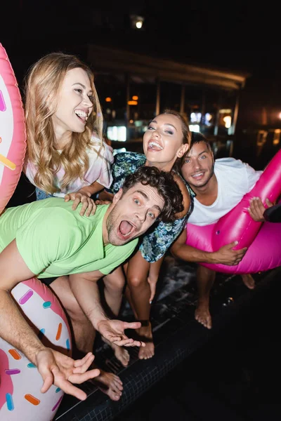 Selective focus of excited young friends having fun with swim rings near pool at night — Stock Photo