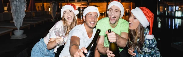 Panoramic shot of excited friends holding bottles of champagne outdoors at night — Stock Photo