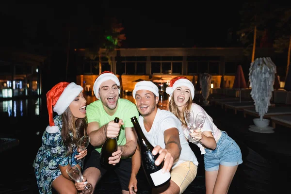 Selective focus of excited friends in santa hats holding bottles of champagne at night outdoors — Stock Photo