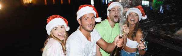 Panoramic orientation of young friends opening bottle of champagne while celebrating new year near near swimming pool at night — Stock Photo