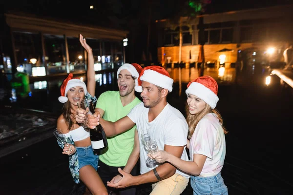 Jóvenes amigos sosteniendo botella de champán y copas mientras celebran el año nuevo cerca de la piscina por la noche - foto de stock