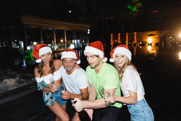 Amici in cappelli di Babbo Natale che tengono bicchieri e bottiglia di champagne vicino alla piscina di notte — Foto stock