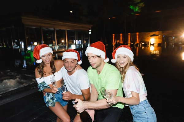 Amigos en sombreros de santa celebración de copas y botella de champán cerca de la piscina por la noche - foto de stock