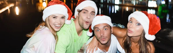 Imagem horizontal de amigos em chapéus de Papai Noel olhando para câmera ao ar livre à noite — Fotografia de Stock