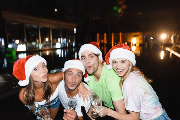 Friends in santa hats embracing while holding champagne and glasses during celebration new year at night outdoors — Stock Photo