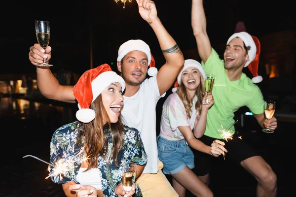 Selective focus of friends in santa hats holding glasses of champagne and sparklers at night — Stock Photo