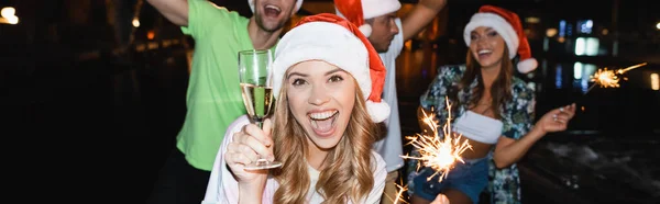 Horizontal image of excited woman holding glass of champagne and sparkler while celebrating new year at night outdoors — Stock Photo