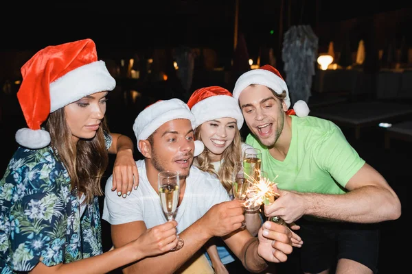 Freunde in Weihnachtsmannmützen mit Champagnergläsern blicken nachts auf Wunderkerzen — Stockfoto