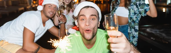 Panoramic shot of man in red santa hat holding sparkler and glass of champagne near friends at night — Stock Photo