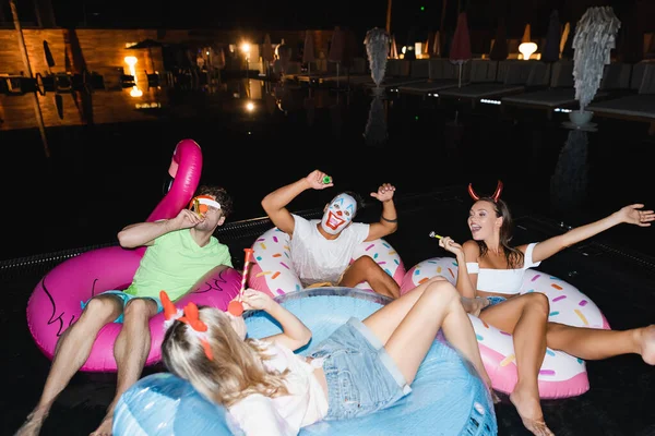 Selective focus of young friends in party headbands and blow ticklers swimming on rings in pool at night — Stock Photo