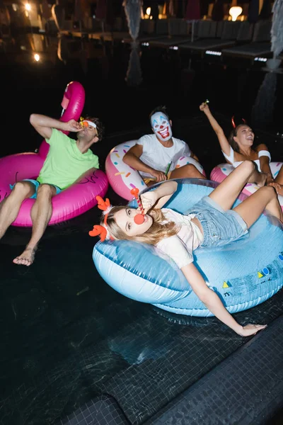 Selective focus of young people in party headbands with blow ticklers swimming on rings in pool during party at night — Stock Photo