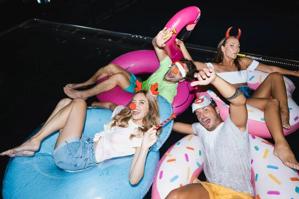 Selektiver Fokus von Freunden mit Partyhörnern, die nachts im Schwimmbad in die Kamera schauen — Stockfoto