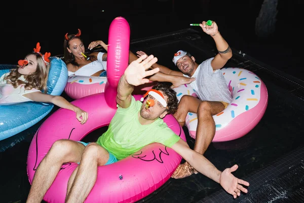 Selektiver Fokus eines Mannes mit Partyhupe, der nachts in der Nähe von Freunden im Schwimmbad in die Kamera winkt — Stockfoto