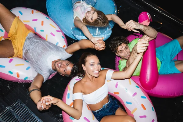 Vista dall'alto di amici che si tengono per mano mentre nuotano sugli anelli in piscina di notte — Foto stock