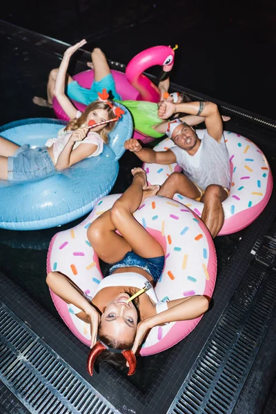 Hochwinkel-Ansicht der Frau in Party-Stirnband mit Schlag Kitzler Schwimmen auf Ring in der Nähe von Freunden in Pool in der Nacht — Stockfoto