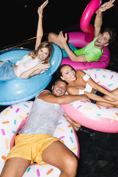 Giovani amici che salutano la fotocamera mentre nuotano sugli anelli in piscina di notte — Foto stock