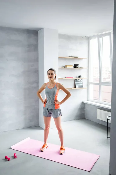 Deportiva de pie con las manos en las caderas en la alfombra de fitness cerca de pesas en casa - foto de stock