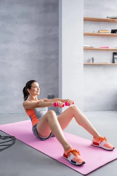 Jovem mulher adulta com halteres fazendo abdominais em esteira de fitness em casa — Fotografia de Stock