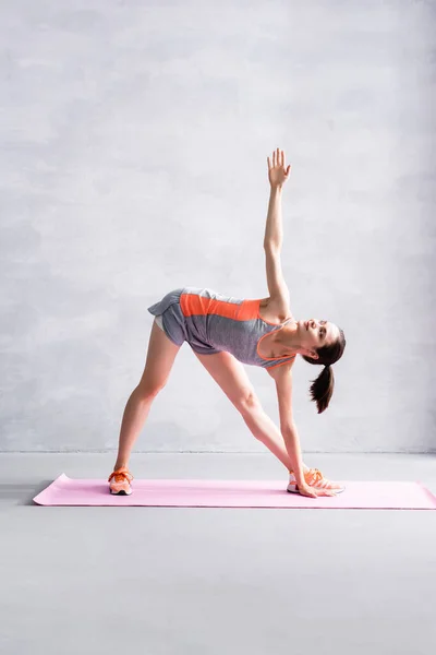 Deportiva con las manos extendidas calentándose en la esterilla de fitness sobre fondo gris - foto de stock