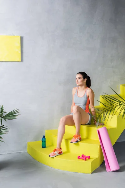Sportlerin sitzt zu Hause auf Treppe neben Flasche, Hanteln und Fitnessmatte — Stockfoto