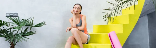 Panoramic orientation of sportswoman sitting on stairs near fitness mat at home — Stock Photo