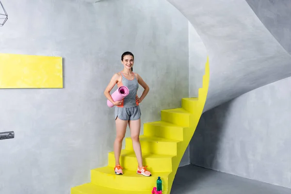 Junge Sportlerin hält Fitnessmatte in der Hand, während sie zu Hause auf der Treppe in die Kamera schaut — Stockfoto