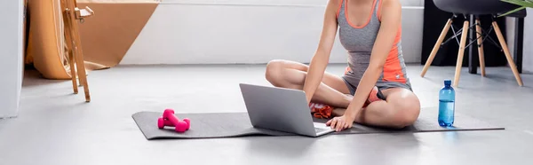 Tiro panorâmico de mulher jovem usando laptop no tapete de fitness em casa — Fotografia de Stock