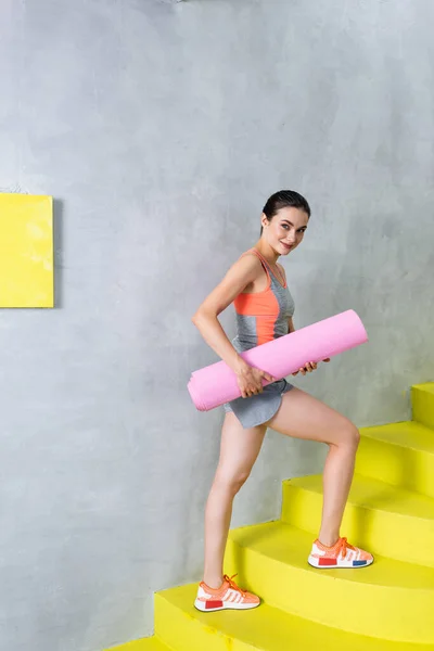 Vista lateral de la deportista con colchoneta de fitness mirando a la cámara en las escaleras en casa - foto de stock