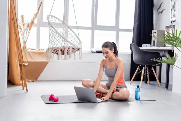Sportiva che utilizza laptop su tappetino fitness vicino a bottiglia d'acqua e manubri — Foto stock
