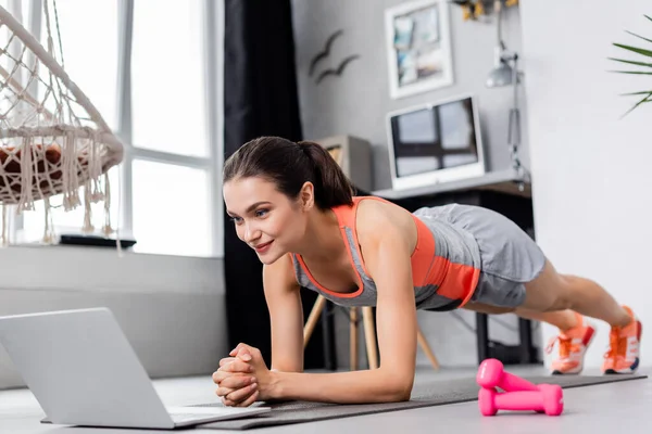 Селективный фокус юной спортсменки, занимающейся доской во время онлайн-тренировок возле гантелей на фитнес-коврике — стоковое фото