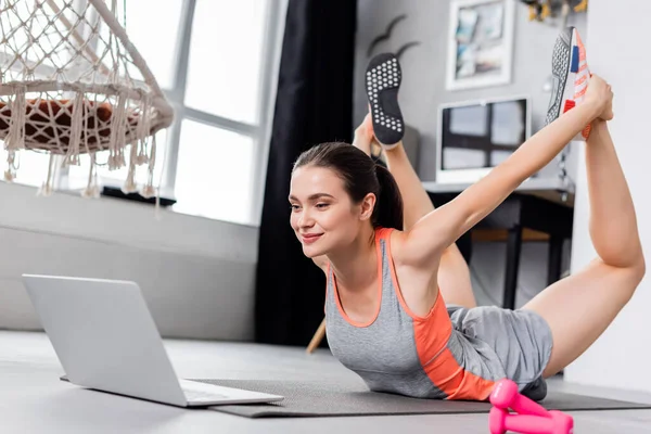 Concentration sélective de la jeune sportive exerçant sur tapis de fitness près d'un ordinateur portable et haltères à la maison — Photo de stock