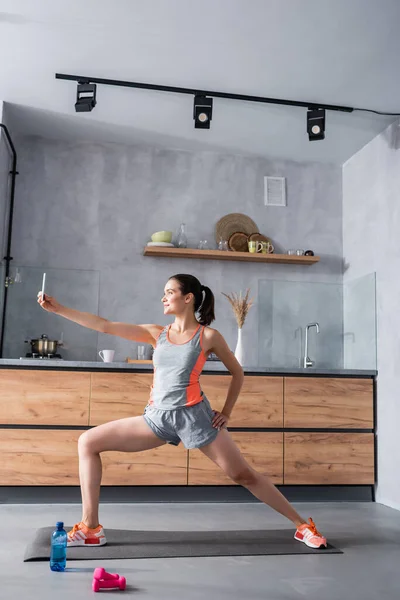 Sportswoman training while holding smartphone near water and dumbbells at home — Stock Photo