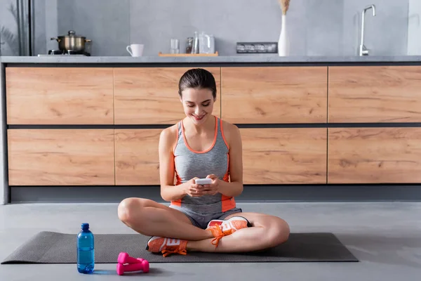 Sportswoman utilisant smartphone sur tapis de fitness près de bouteille d'eau et haltères dans la cuisine — Photo de stock