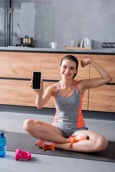 Sportswoman montrant smartphone avec écran vierge près de bouteille d'eau et d'équipement sportif dans la cuisine — Photo de stock