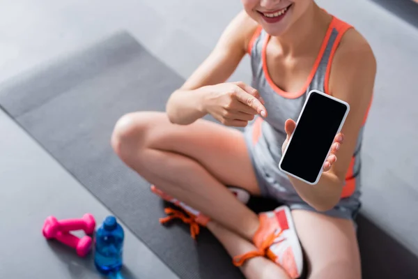 Обрізаний вид жінки в спортивному одязі, що вказує на смартфон з порожнім екраном на фітнес-маті — стокове фото