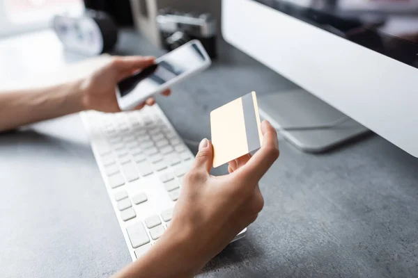 Vista recortada del teletrabajador con tarjeta de crédito y teléfono inteligente cerca de la computadora en la mesa en casa - foto de stock