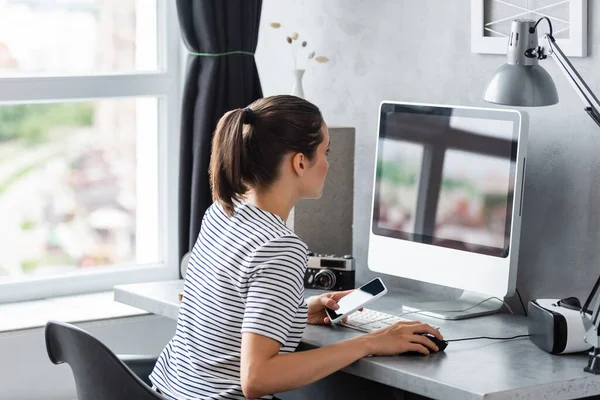 Joven freelancer sosteniendo el teléfono inteligente mientras usa el ordenador en casa - foto de stock