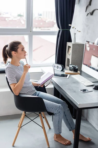 Barfüßiger Freiberufler hält Stift und Notizbuch in der Nähe von Gadgets auf dem Tisch — Stockfoto