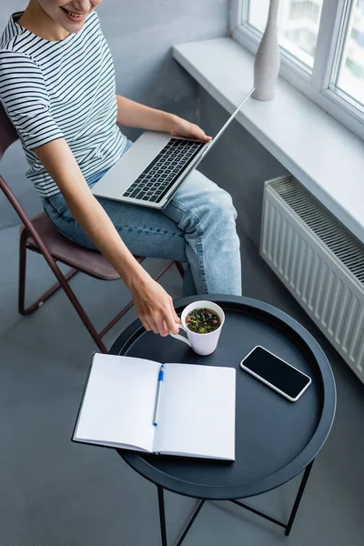 Vista recortada del freelancer sosteniendo la taza de café mientras usa el portátil cerca del teléfono inteligente y el portátil en casa - foto de stock