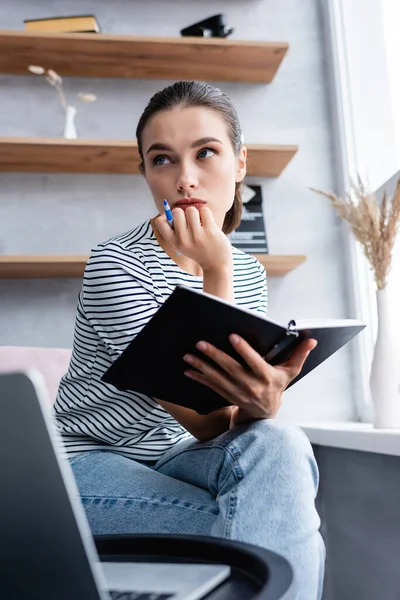 Selective focus of pensive freelancer holding pen and notebook near laptop at home — Stock Photo