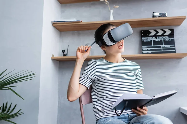 Selective focus of pensive woman in virtual reality headset holding pen and notebook at home — Stock Photo