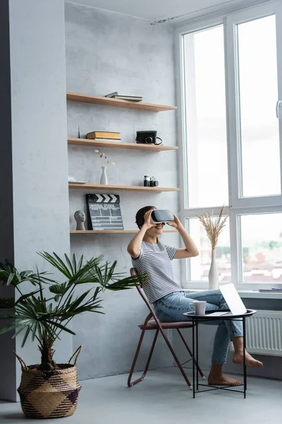 Freelancer descalzo usando auriculares vr cerca de la computadora portátil y la taza en la mesa de café en casa - foto de stock